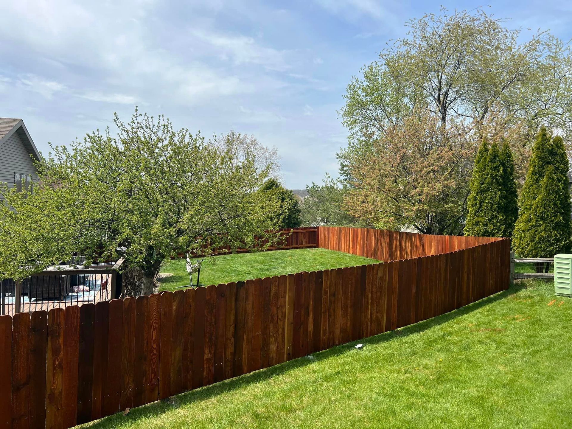Brown wooden fence surrounds a green lawn with trees under a blue sky.