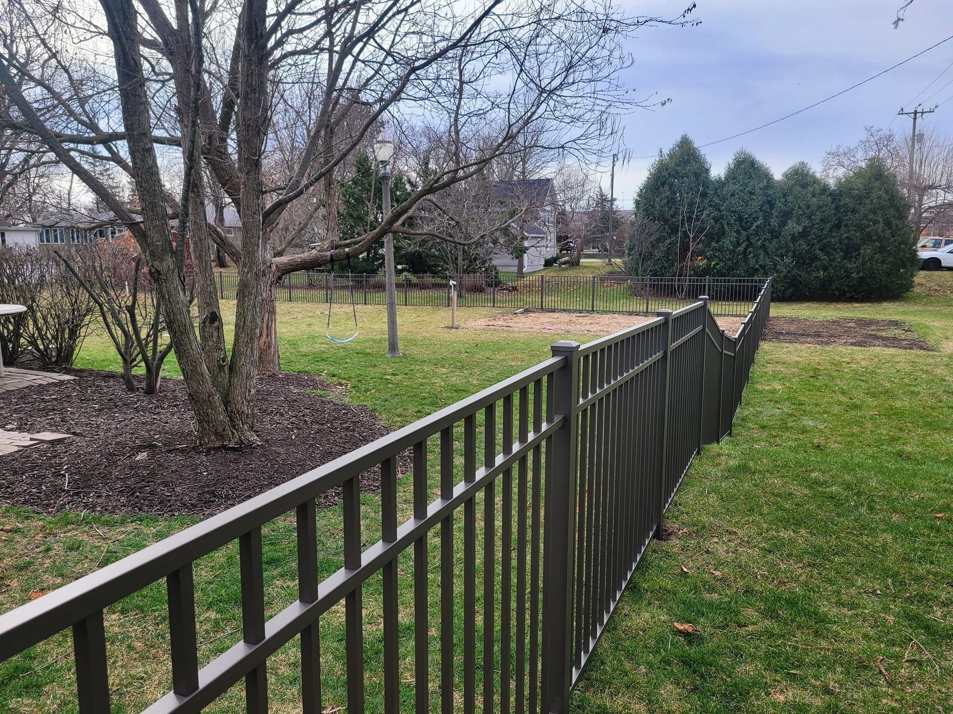 Brown metal fence on grassy lawn, trees and houses in the background. Cloudy sky.