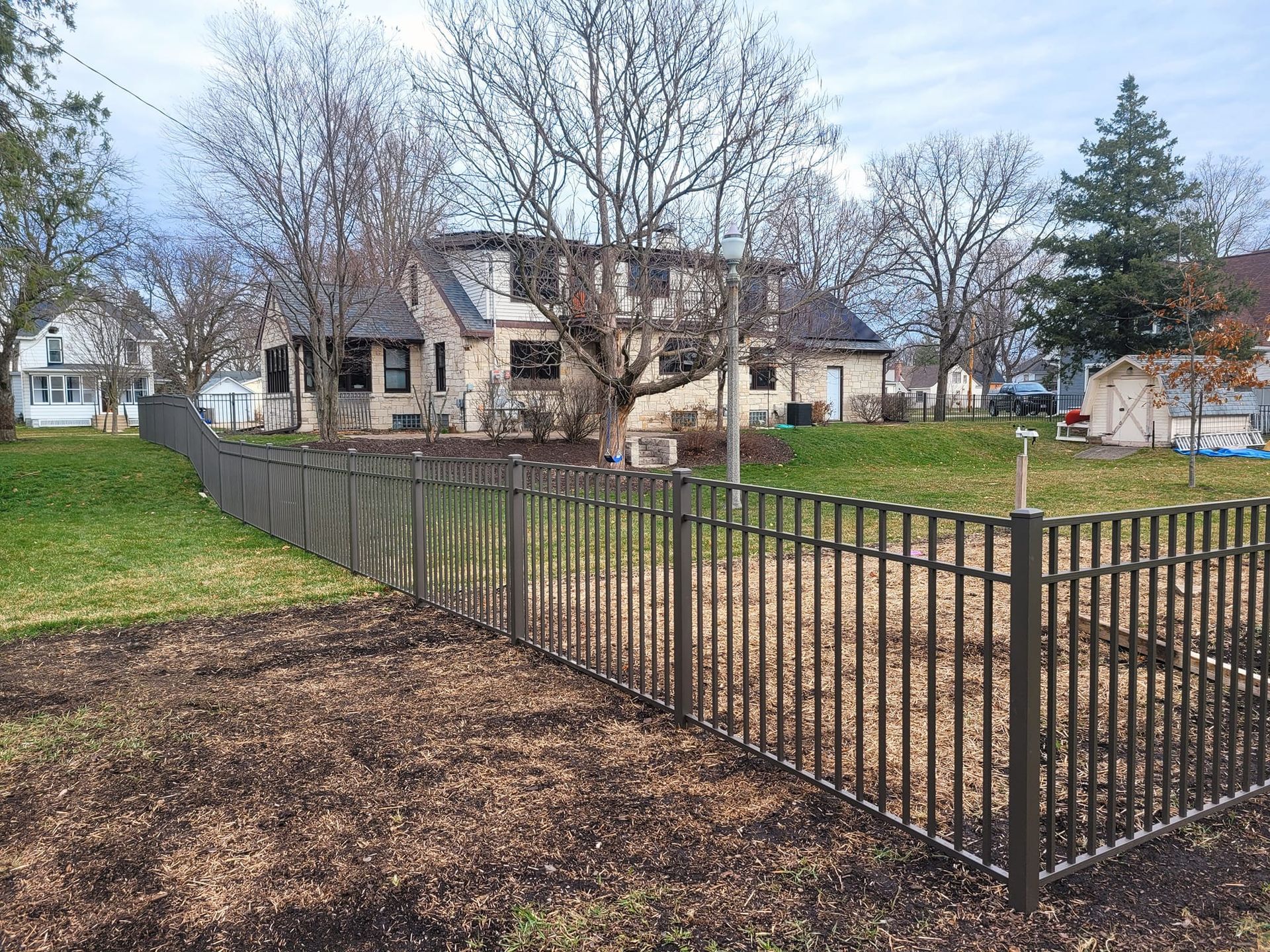 Brown metal fence encloses a brown patch of land in front of a two-story home with a cloudy sky background.