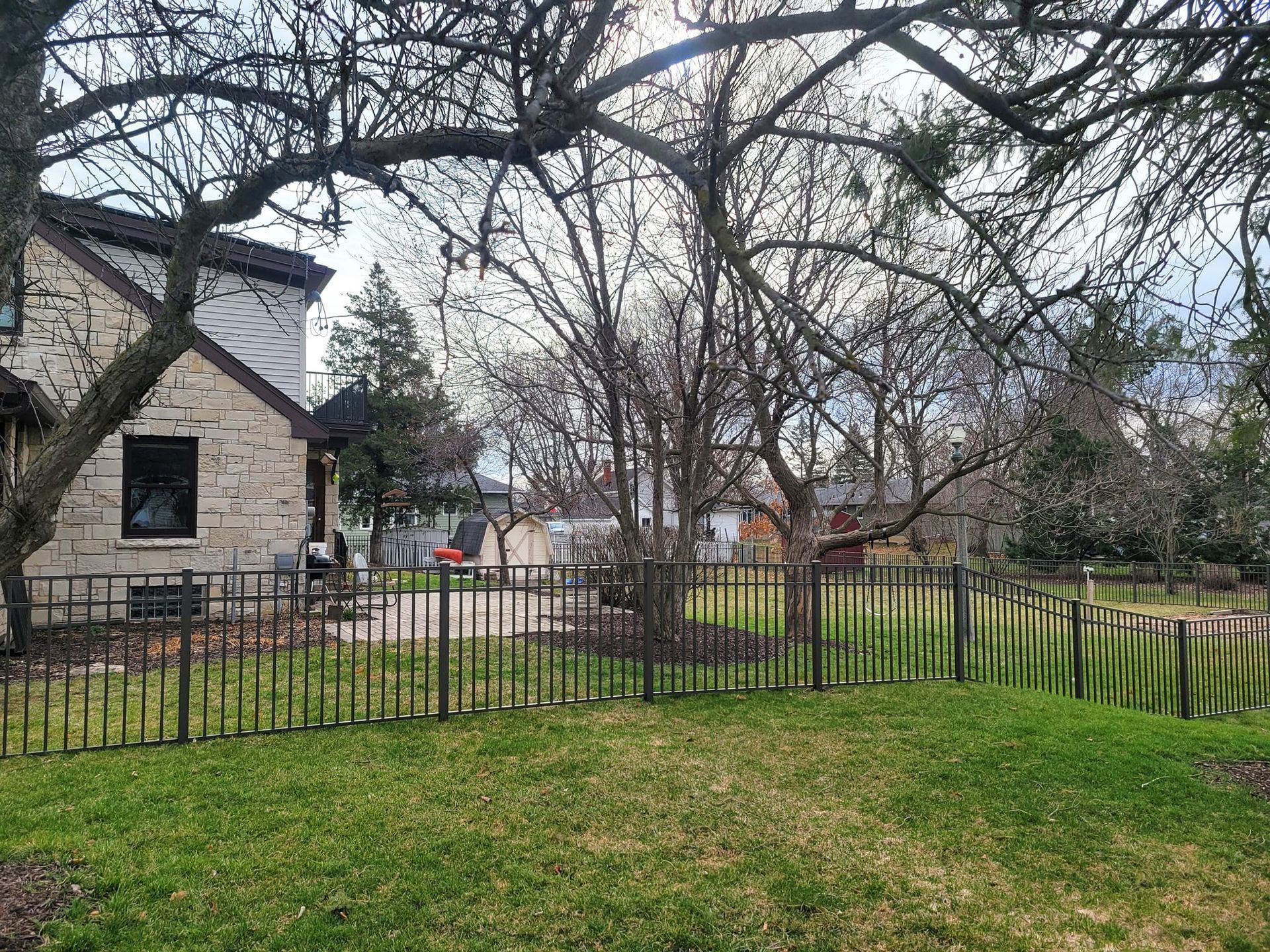 Backyard with a black metal fence, brick house, and bare trees against a cloudy sky.