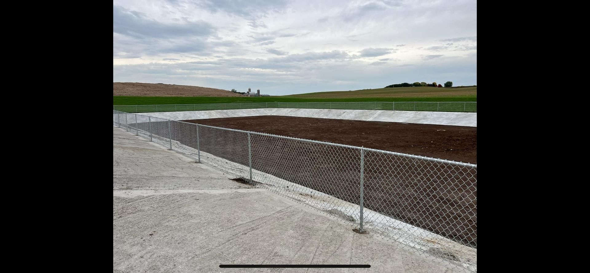 Chain-link fence along a gravel path, bordering a brown field and flooded area, with a cloudy sky.