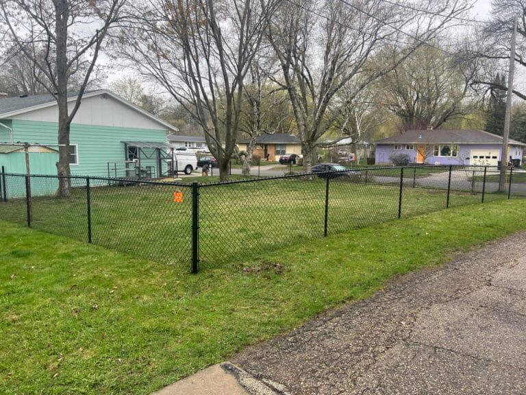 Black chain-link fence surrounds a grassy yard in a residential neighborhood with houses and trees in the background.