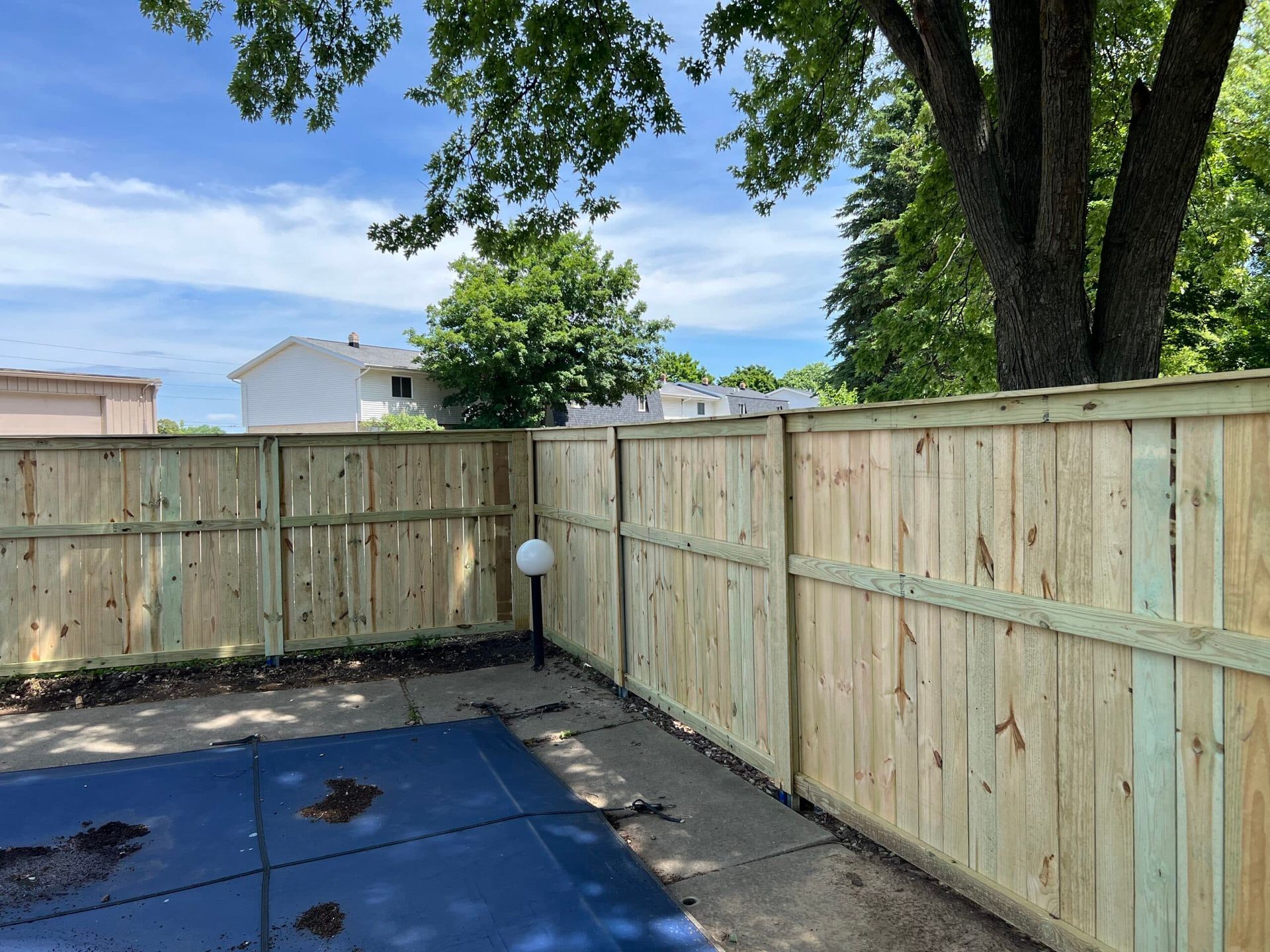 Wooden fence surrounds a backyard with a blue pool cover and trees, under a partly cloudy sky.