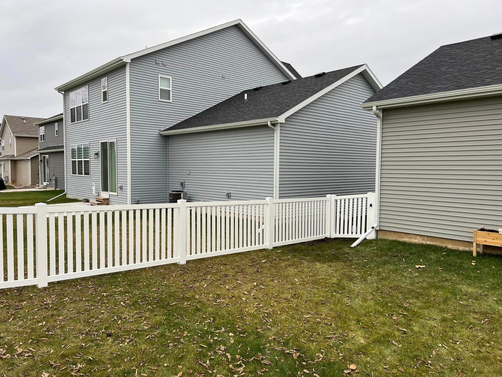 Backyard with white picket fence, gray siding house, and overcast sky.