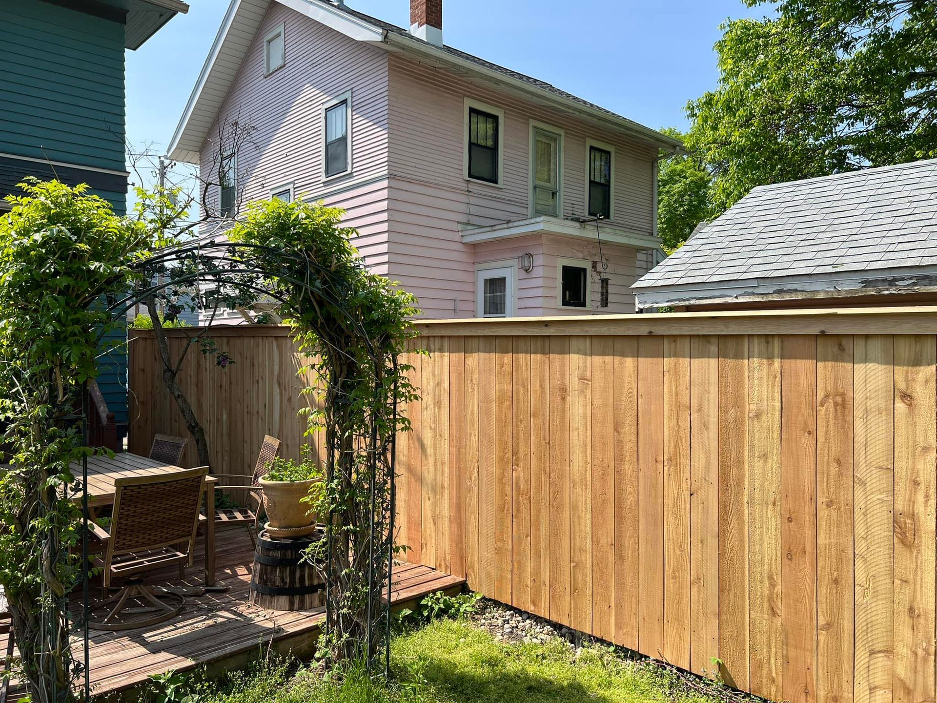 Wooden fence and arbor with climbing plants in front of a pink house and a house with a gray roof.