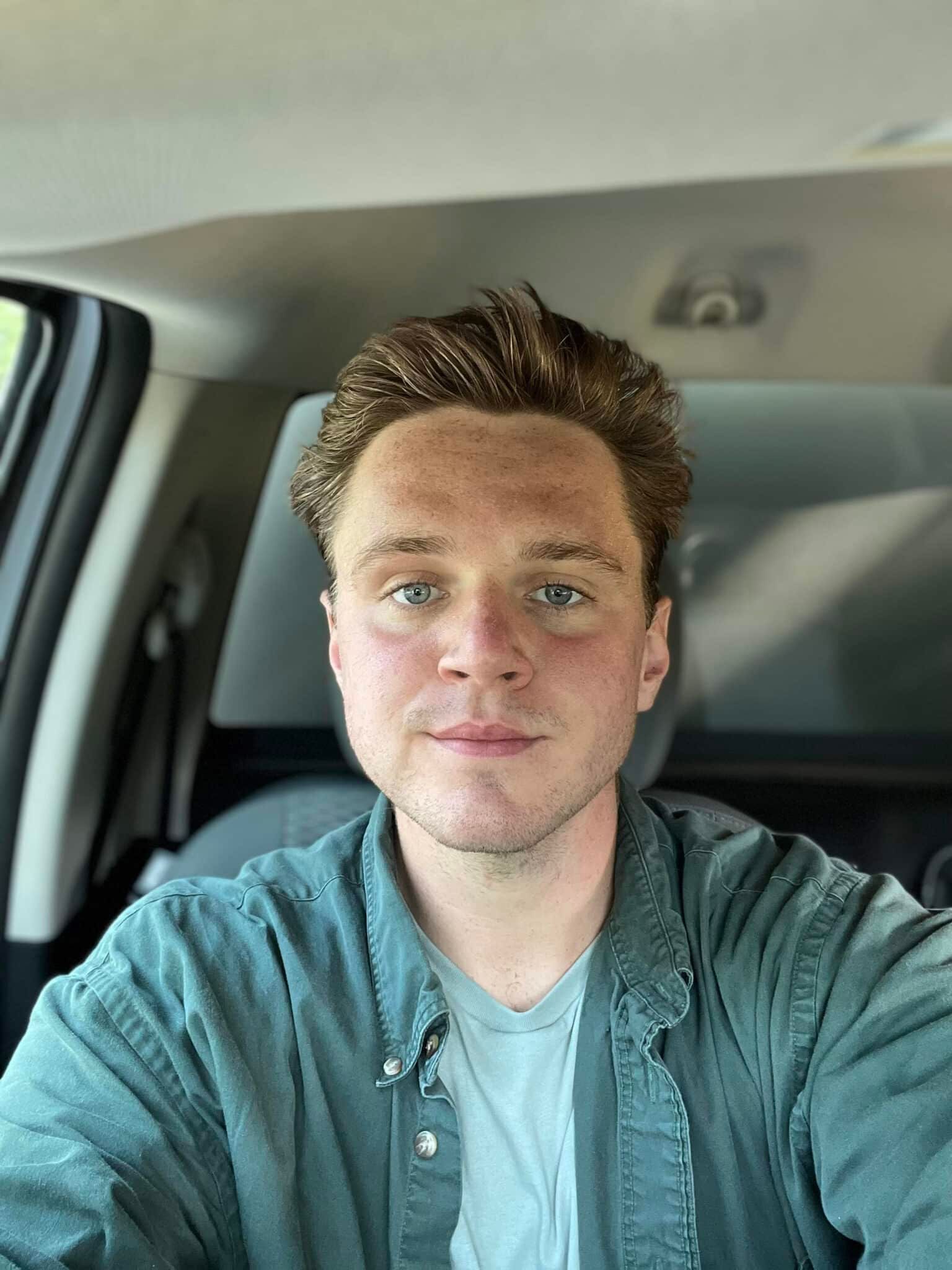Man in car, wearing green shirt, looking at camera with a neutral expression.