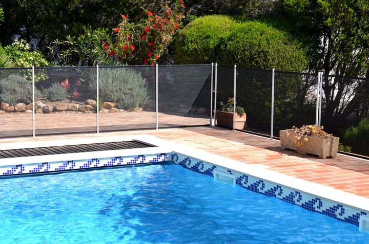Pool with safety fence, surrounded by brick patio and lush greenery.
