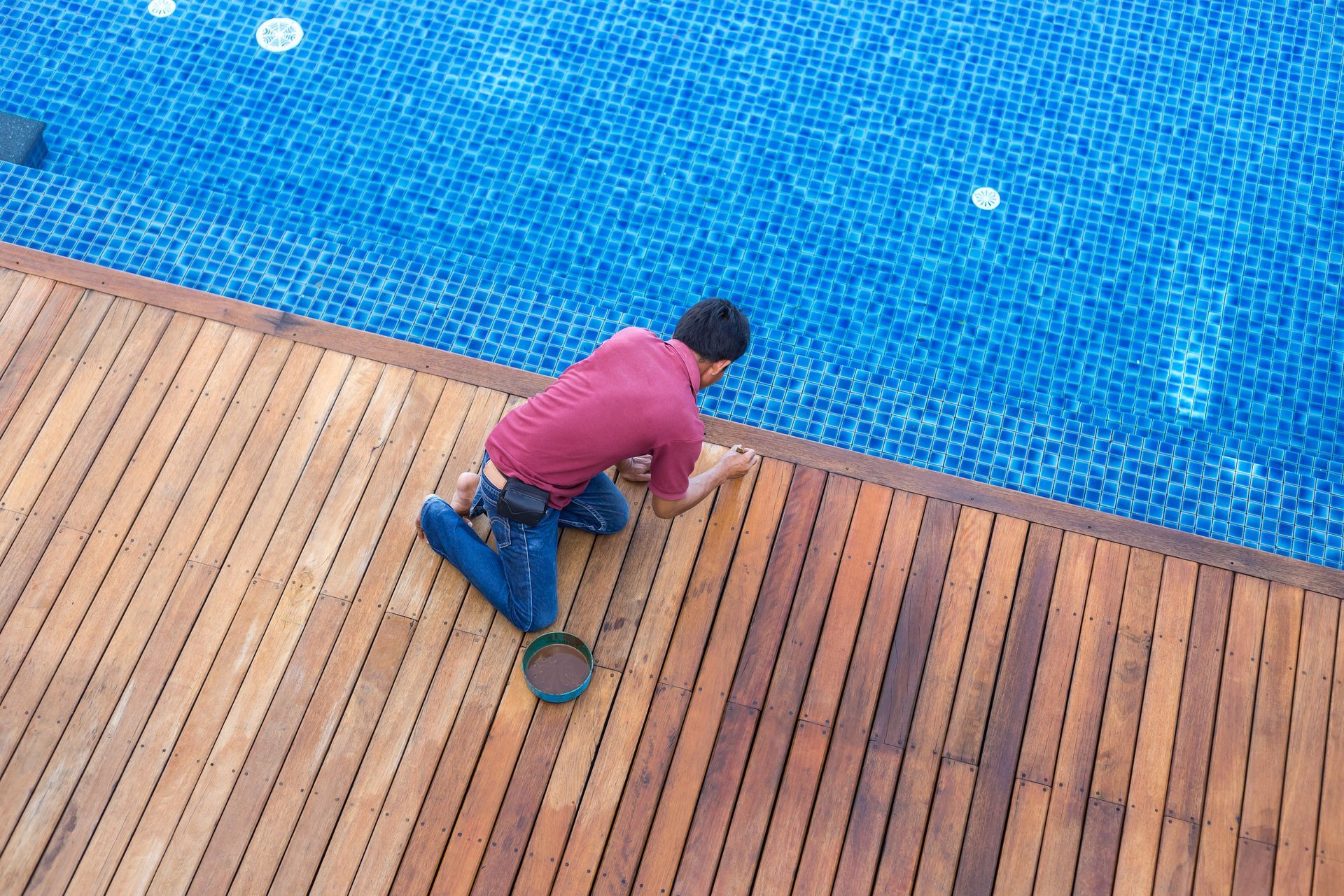 Man staining a wooden deck next to a blue swimming pool.