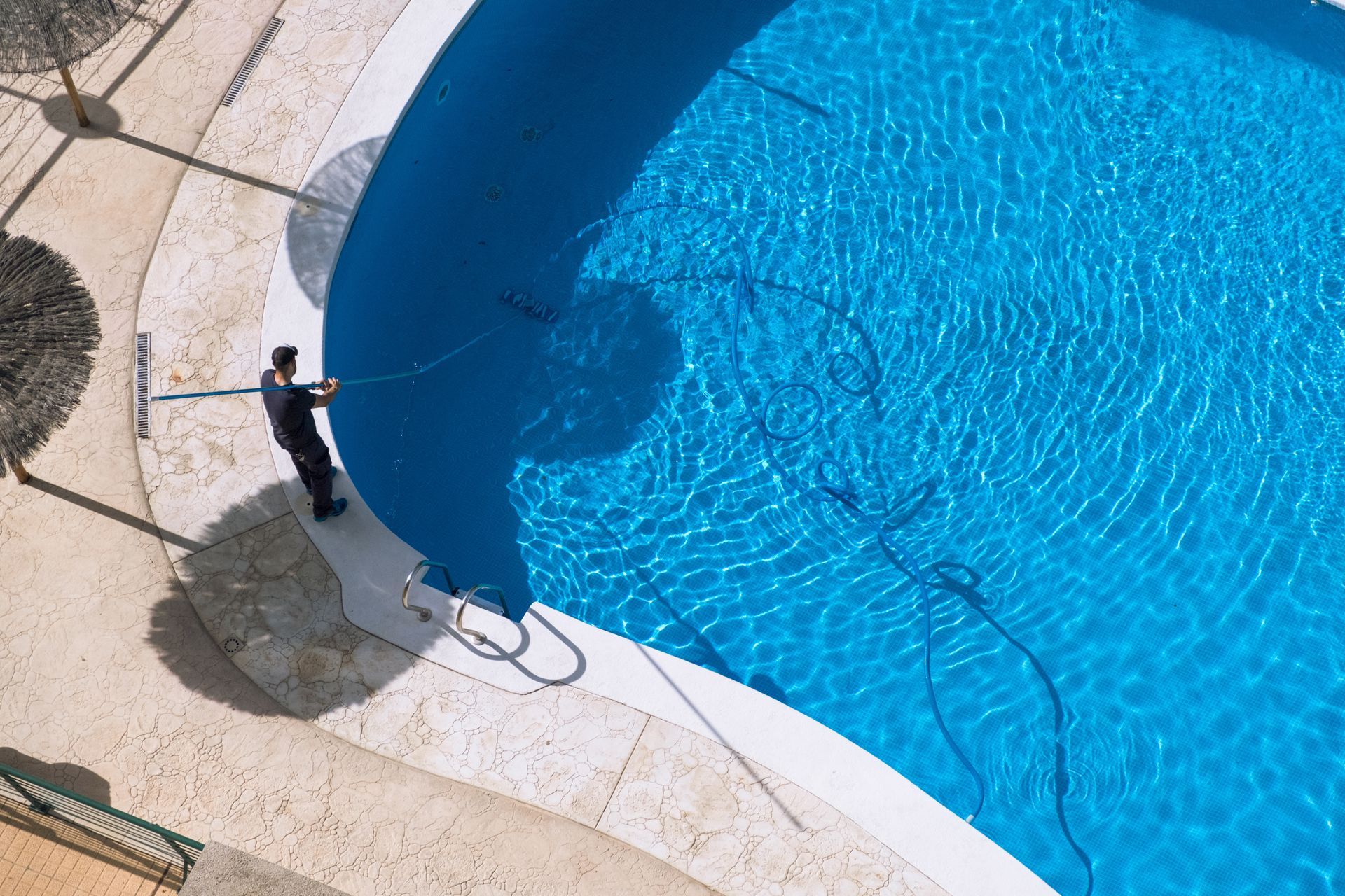 A person cleans a blue, kidney-shaped pool with a hose on a sunny day.