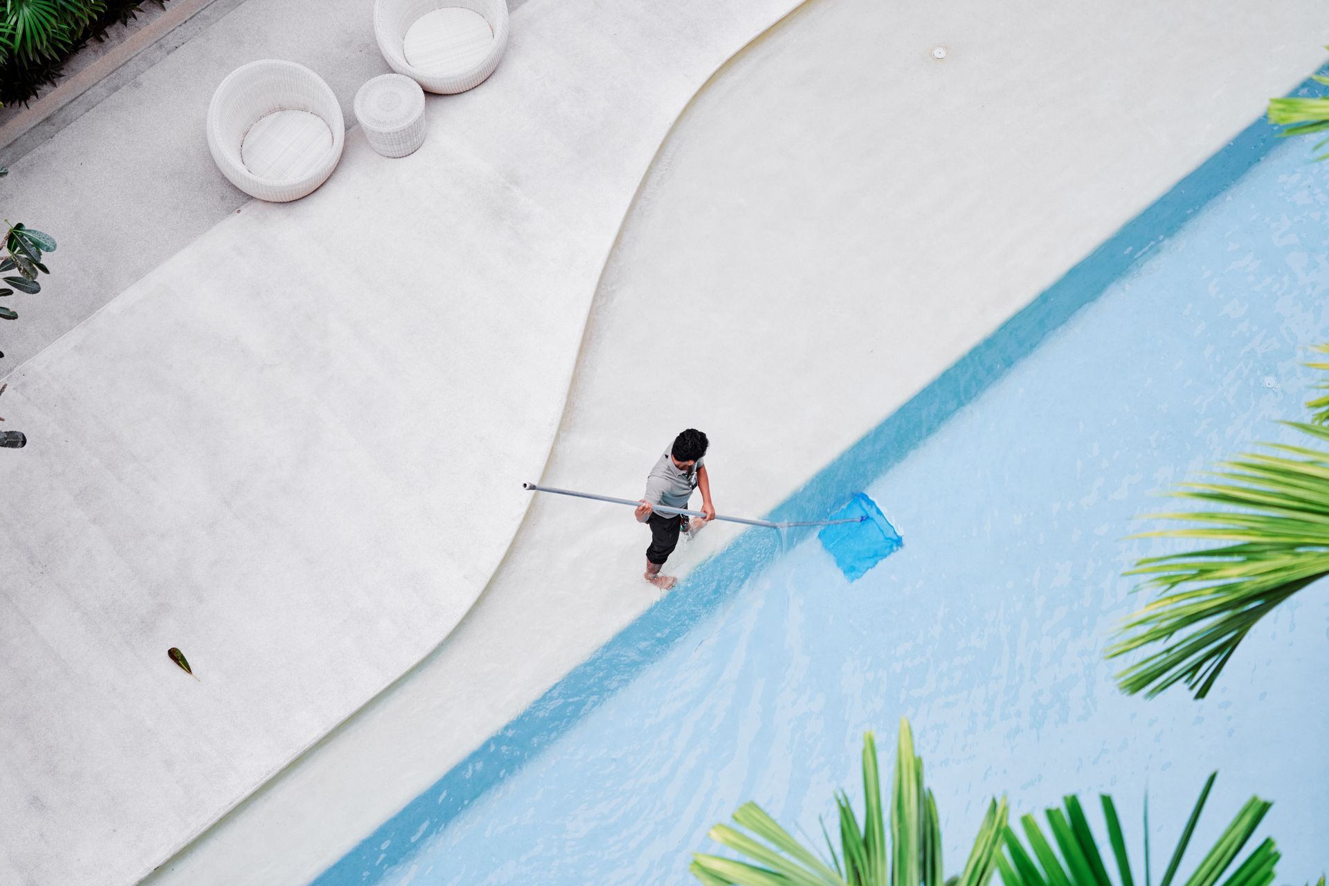 Person cleaning a blue swimming pool with a net; white patio with chairs and palm trees.