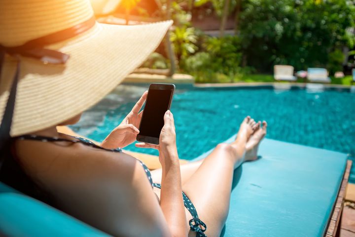 Woman in swimsuit and hat using phone poolside in the sun.