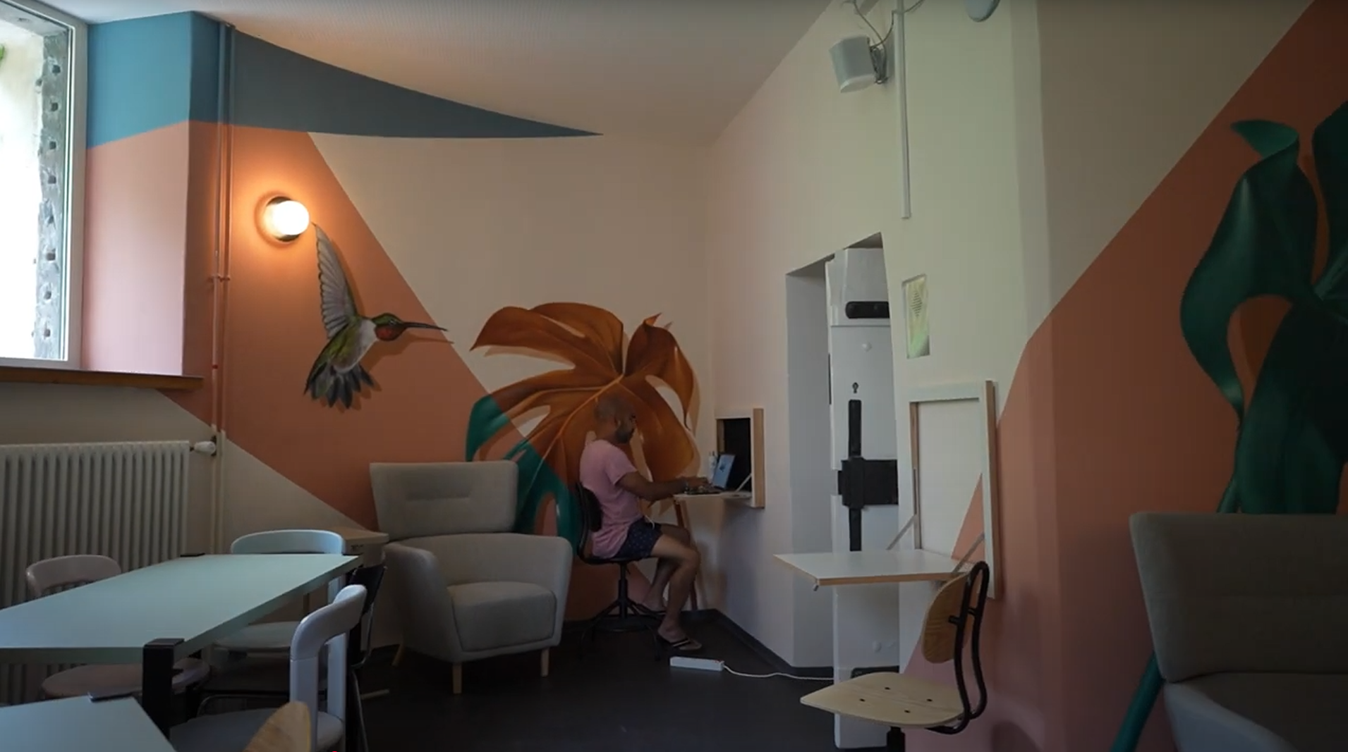 A room with painted murals, chairs, and tables. Two people are interacting with a computer near a floral mural.