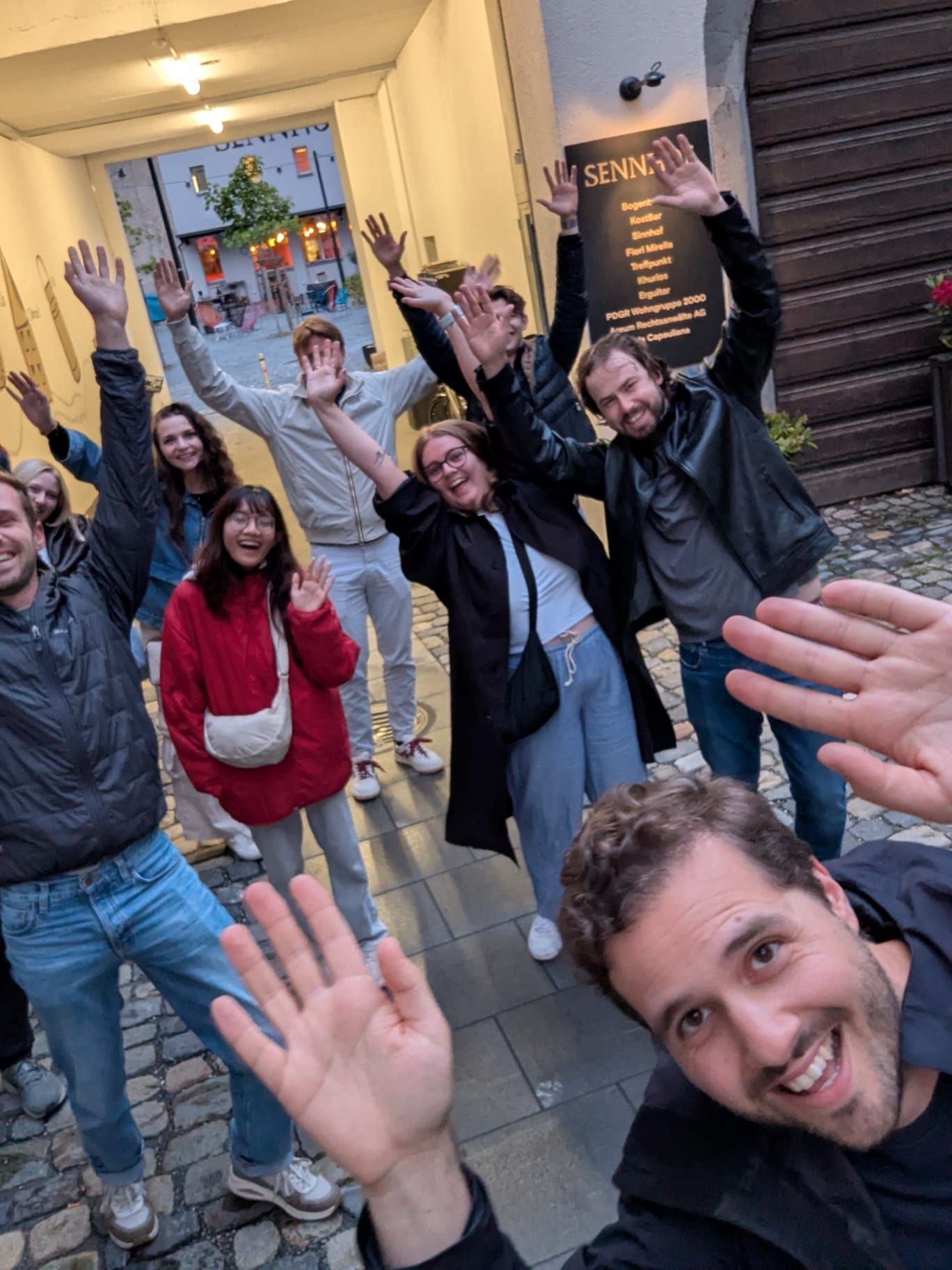 Eine Gruppe von Menschen winkt und lächelt in einem schmalen Gehweg, im Hintergrund ein Backsteingebäude, einige tragen Jacken.