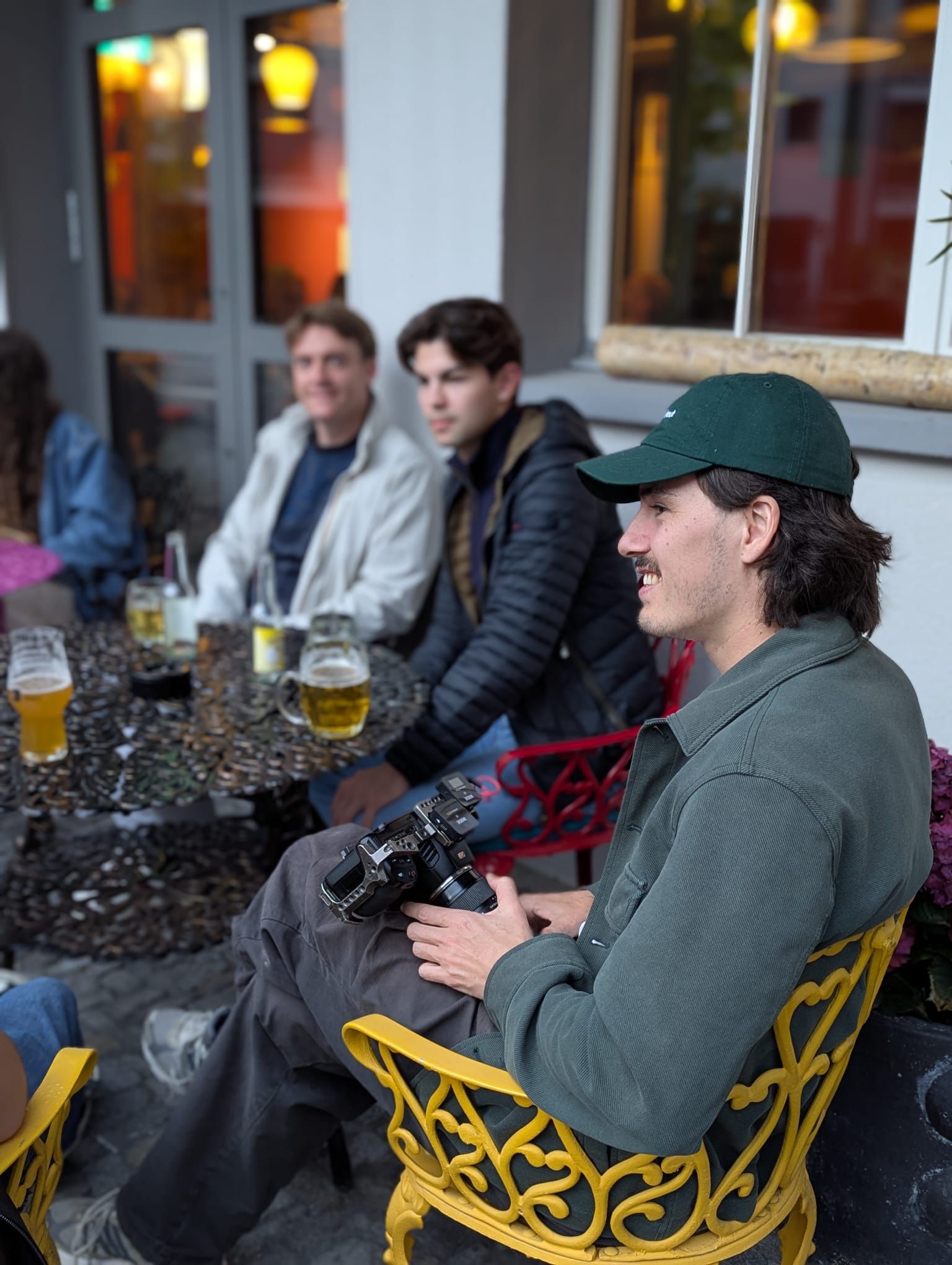 Eine Gruppe junger Männer sitzt an einem Tisch im Freien. Einer hält eine Kamera in der Hand und trägt eine grüne Kappe. Auf dem Tisch steht Bier.