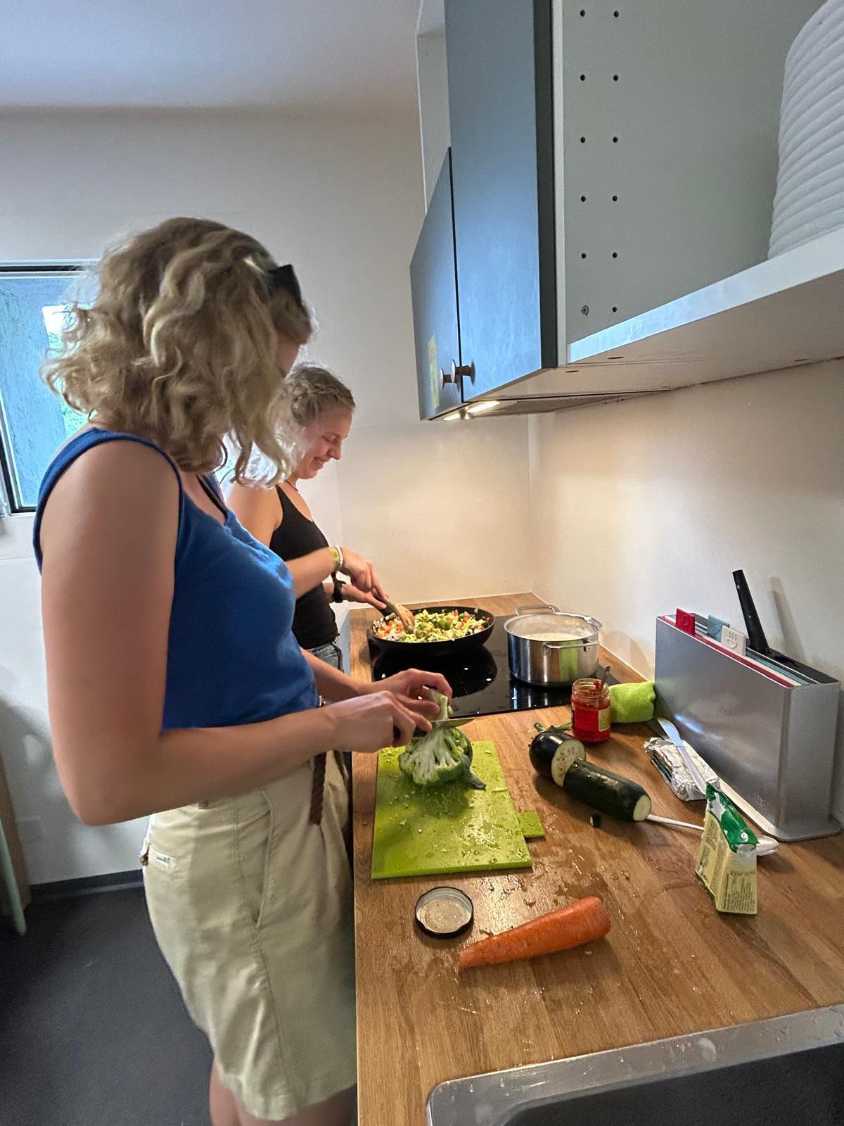 Zwei Frauen kochen in einer Küche. Eine schneidet Gemüse, die andere rührt das Essen in einer Pfanne auf dem Herd um.