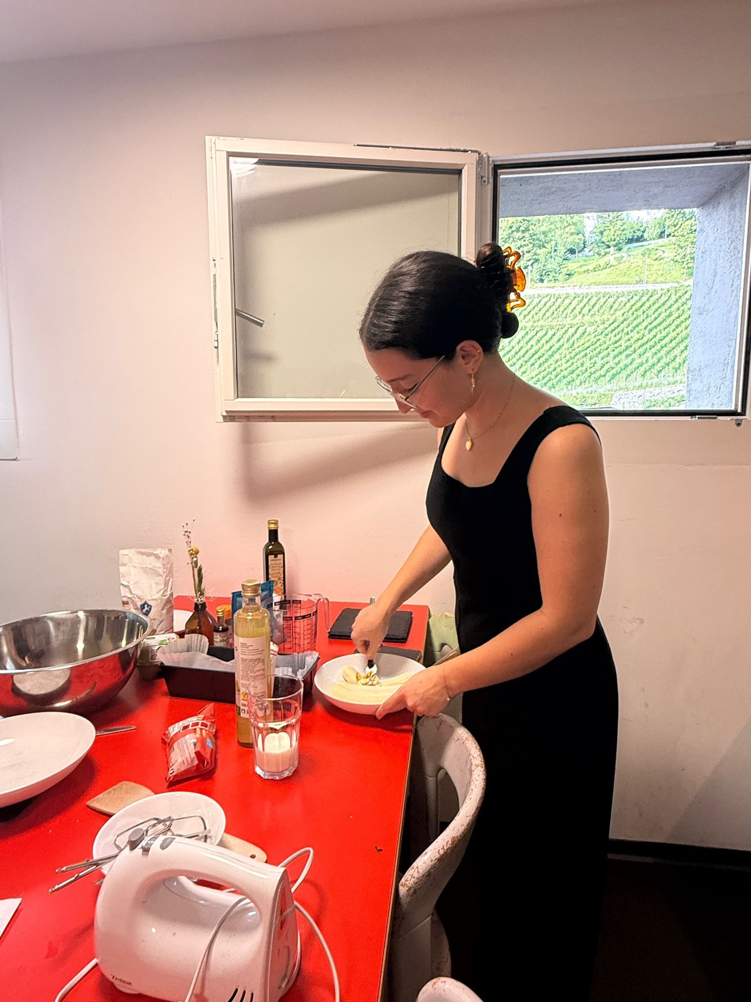 Frau im schwarzen Kleid kocht am roten Tisch mit Zutaten, in der Nähe des Fensters.