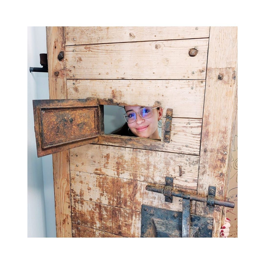 Eine Frau mit Brille späht lächelnd durch ein kleines Fenster in einer alten Holztür.