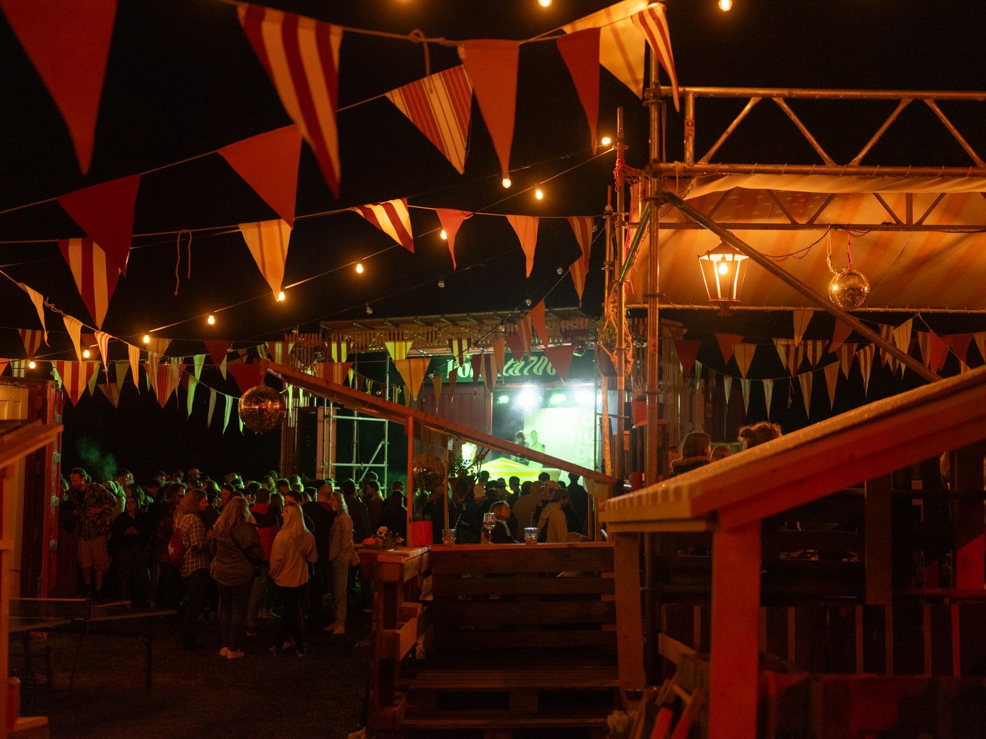 Nighttime outdoor festival with colorful pennant flags, crowds, and bright stage lights under a canopy