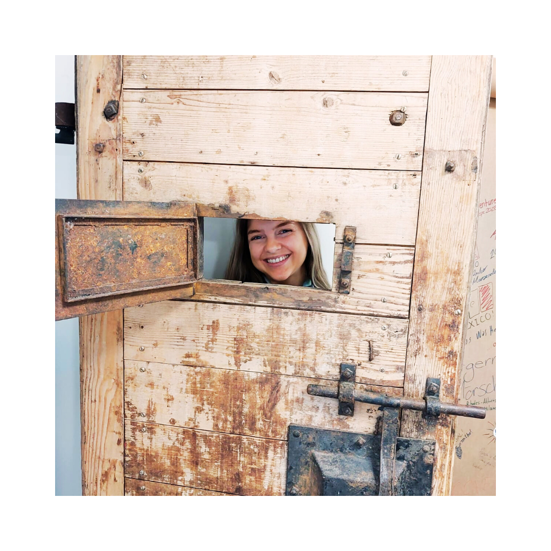 Eine Frau späht lächelnd durch eine kleine Öffnung in einer alten, verwitterten Holztür.
