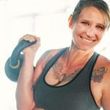 A woman is holding a kettlebell on her shoulder and smiling.