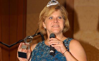 A woman wearing a tiara is holding a microphone and a trophy.
