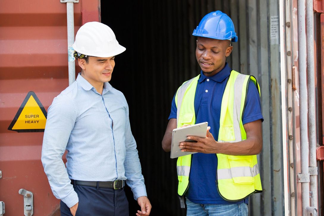 two men are standing next to each other in front of a shipping container looking at a tablet