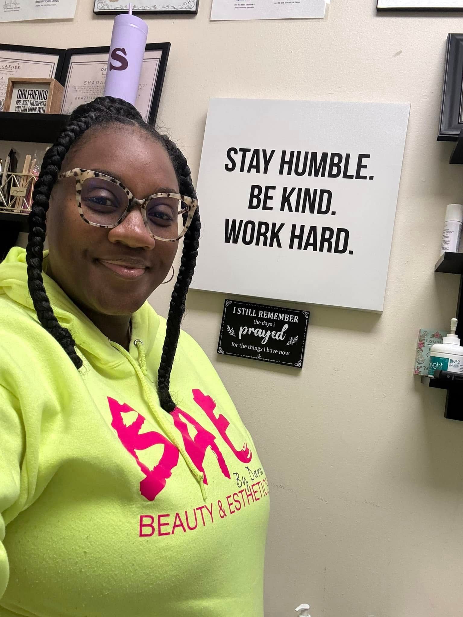 A woman in a neon yellow hoodie is standing in front of a sign that says `` stay humble be kind work hard ''.