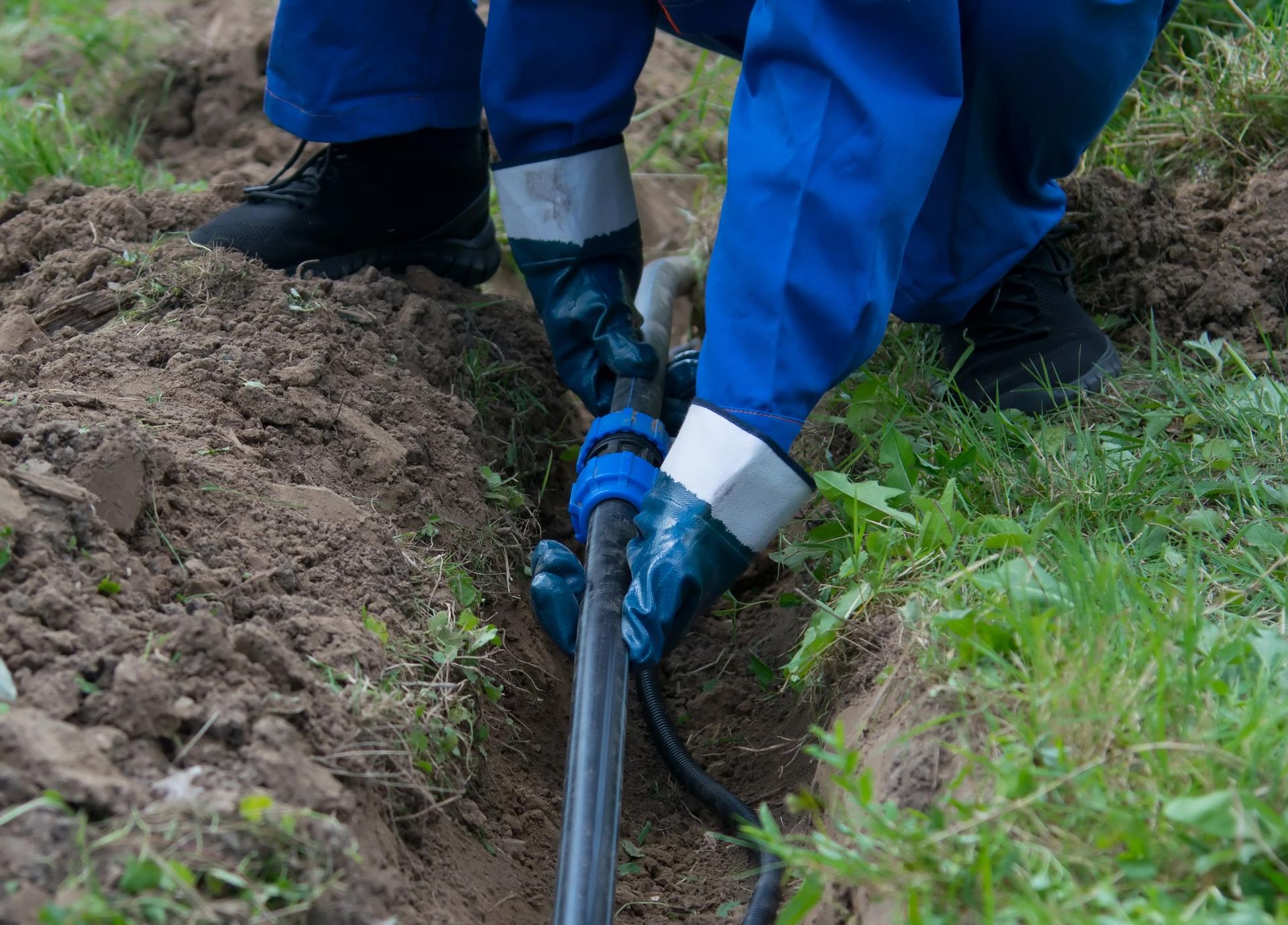 Person in blue work suit connecting black pipes in a trench outdoors.