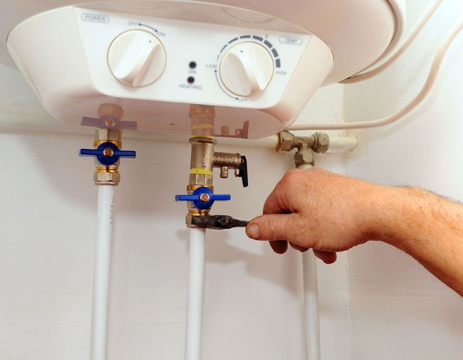 Hand using a wrench to adjust a valve connected to a water heater; white pipes, blue valves.