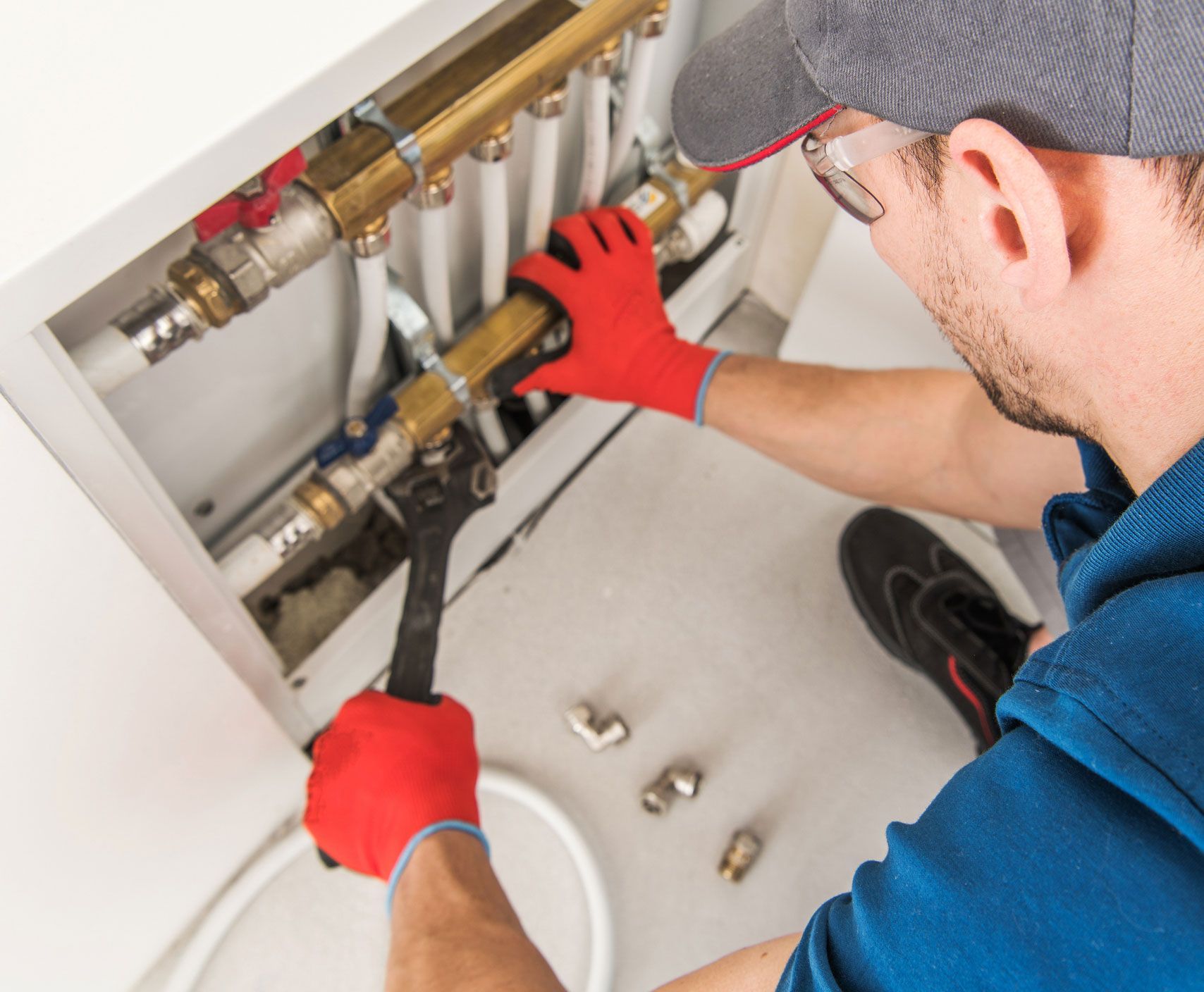 Plumber in red gloves using wrench on brass pipes in a white cabinet.