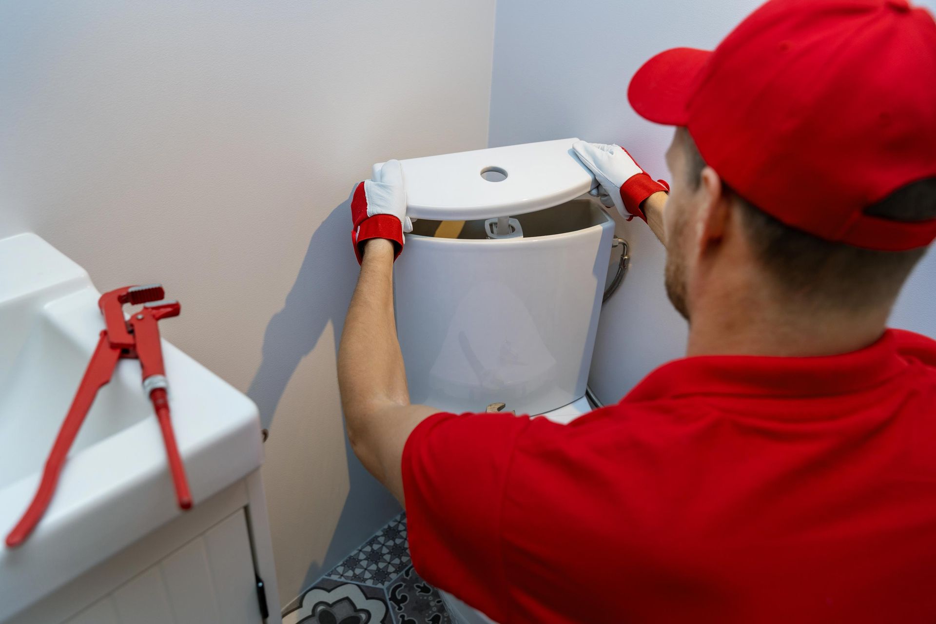 Plumber in red uniform installs toilet tank in a bathroom, wrench on the sink.