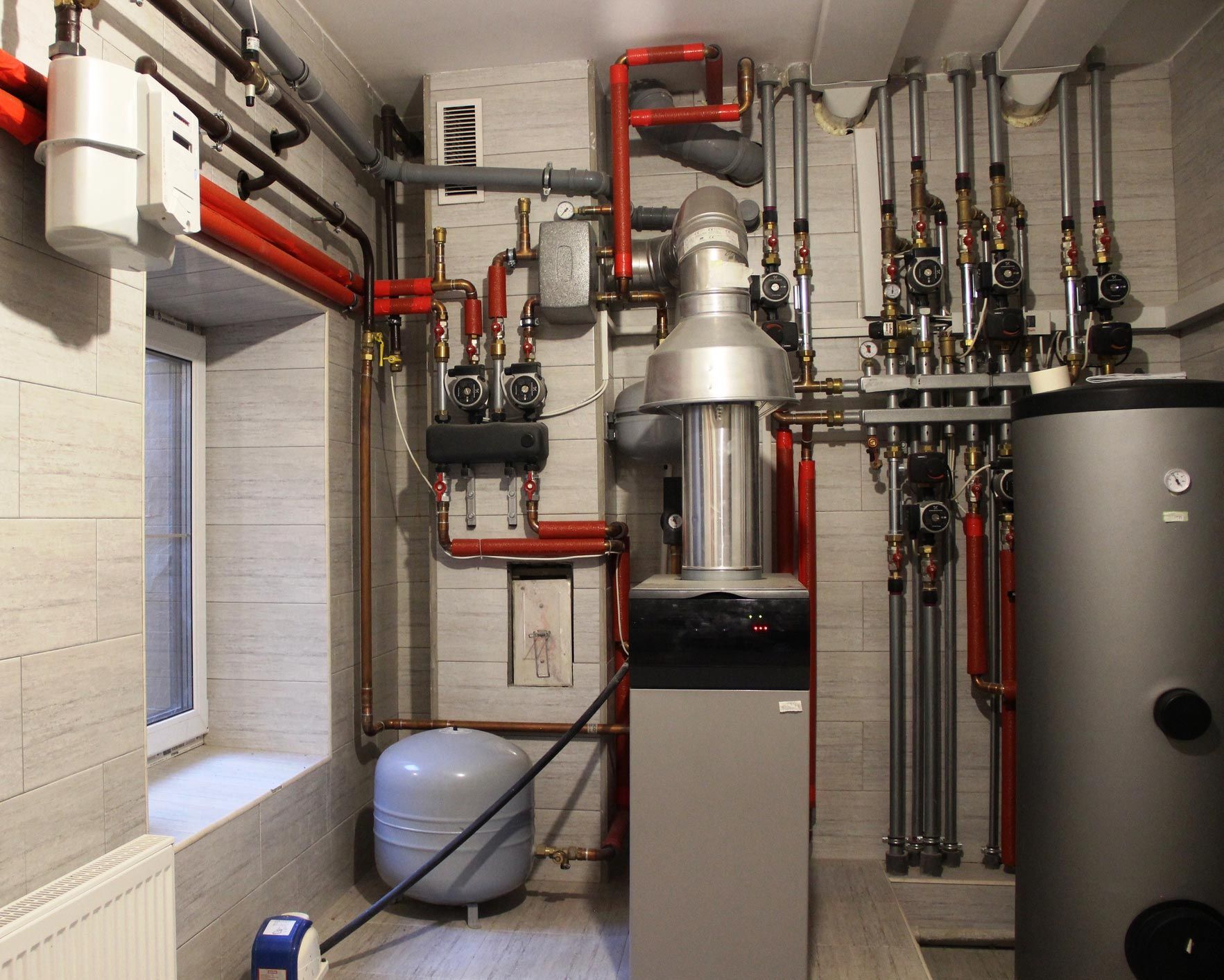 A boiler room with complex heating system pipes and tanks, with a gas meter and small window.
