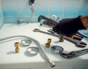 Plumber's hand in black glove holding wrench over tools and plumbing parts on a white surface.