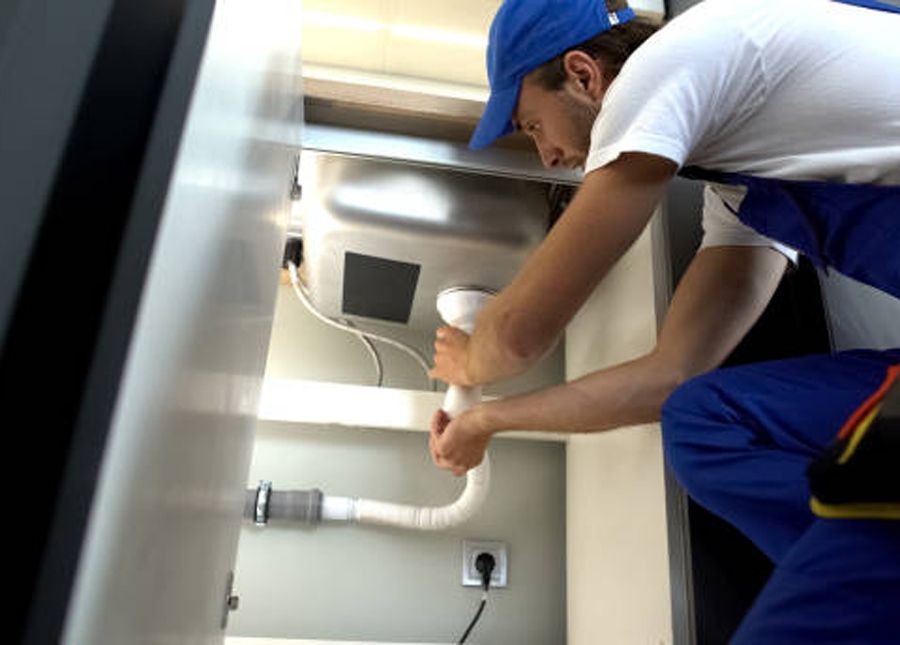 Plumber in blue overalls and cap working on pipes under a kitchen sink.