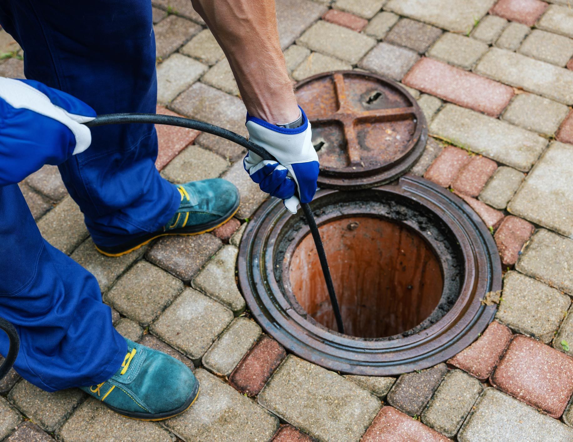 Man using a drain snake, clearing a pipe in a brick-paved area.