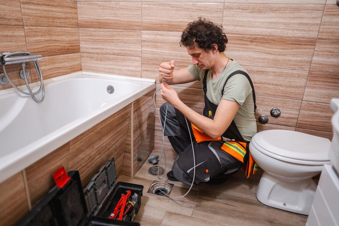 Plumber fixing bathtub drain in a bathroom. Man kneeling, holding tools, near tub and toilet.
