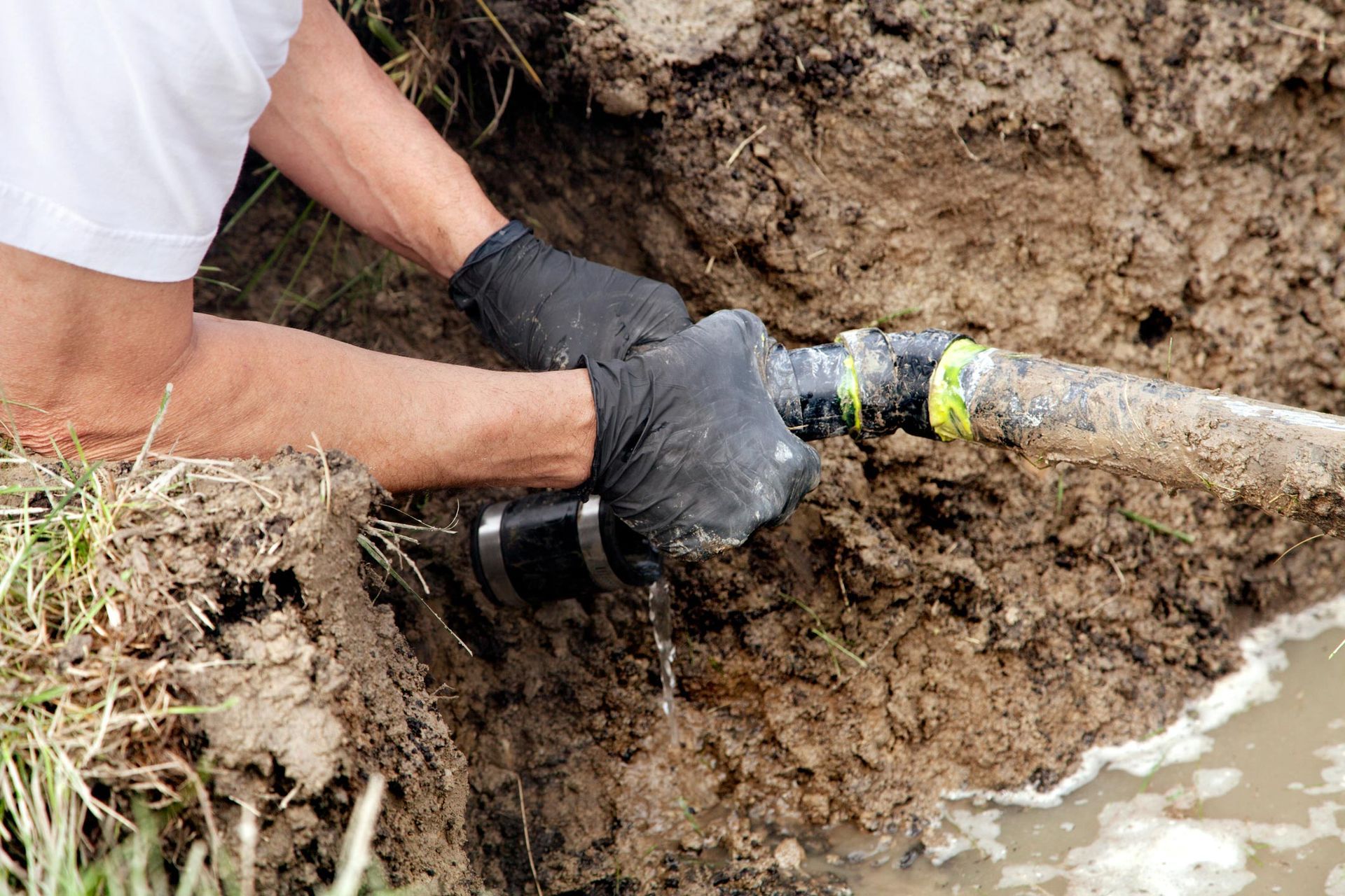 Hands in black gloves fix a broken pipe in a dirt trench, water leaking.
