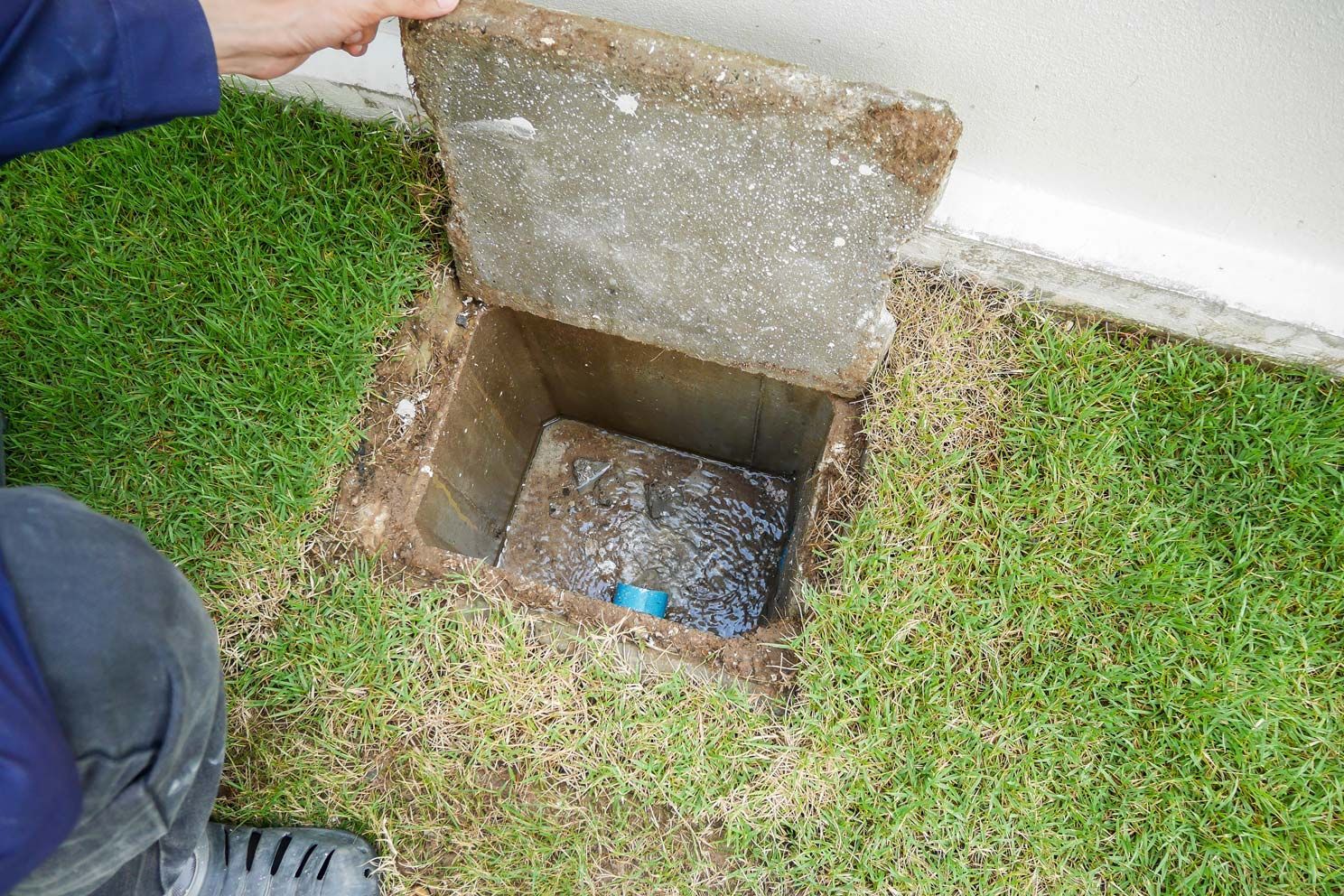 A hand lifting a concrete cover off a rectangular ground access box in green grass.