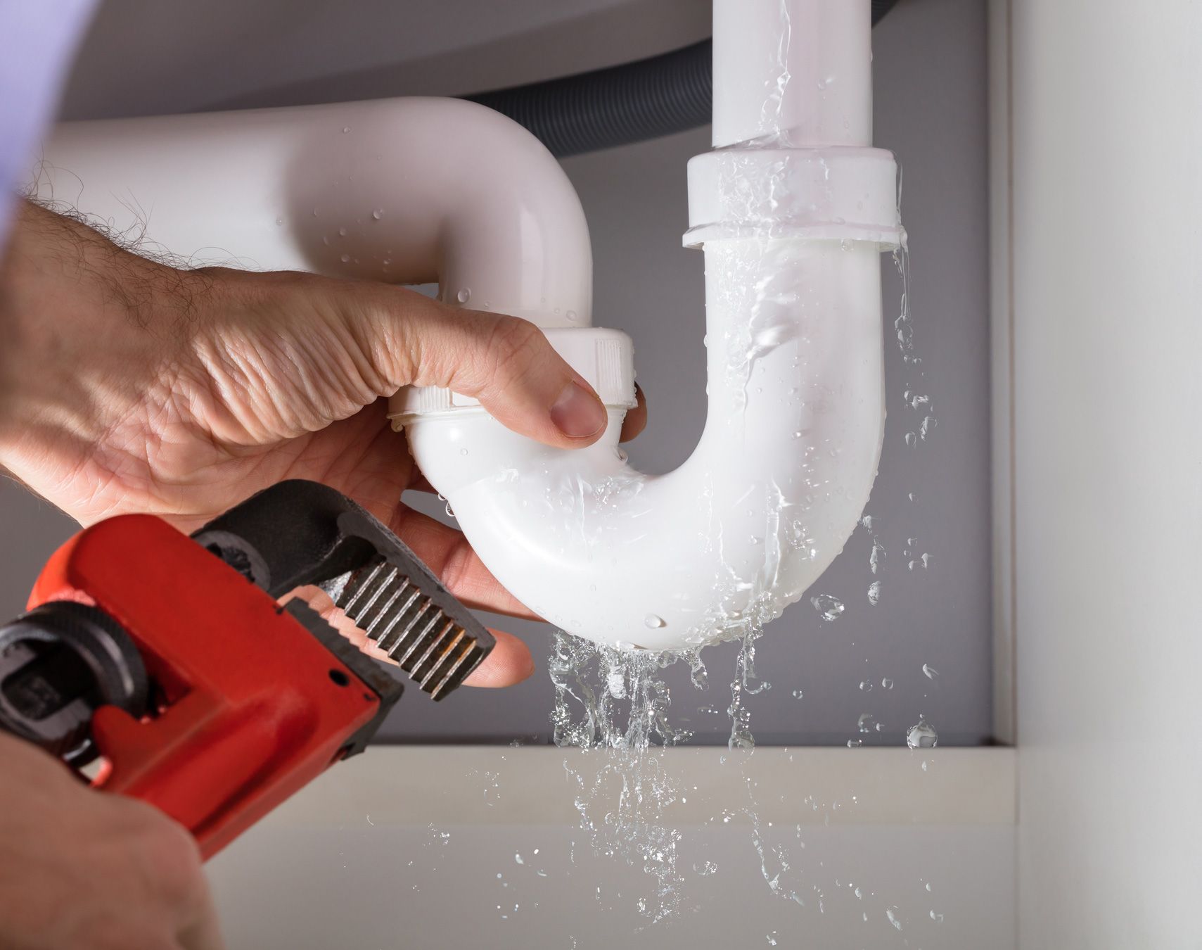 Plumber using a wrench to fix a leaking white pipe under a sink. Water is dripping.
