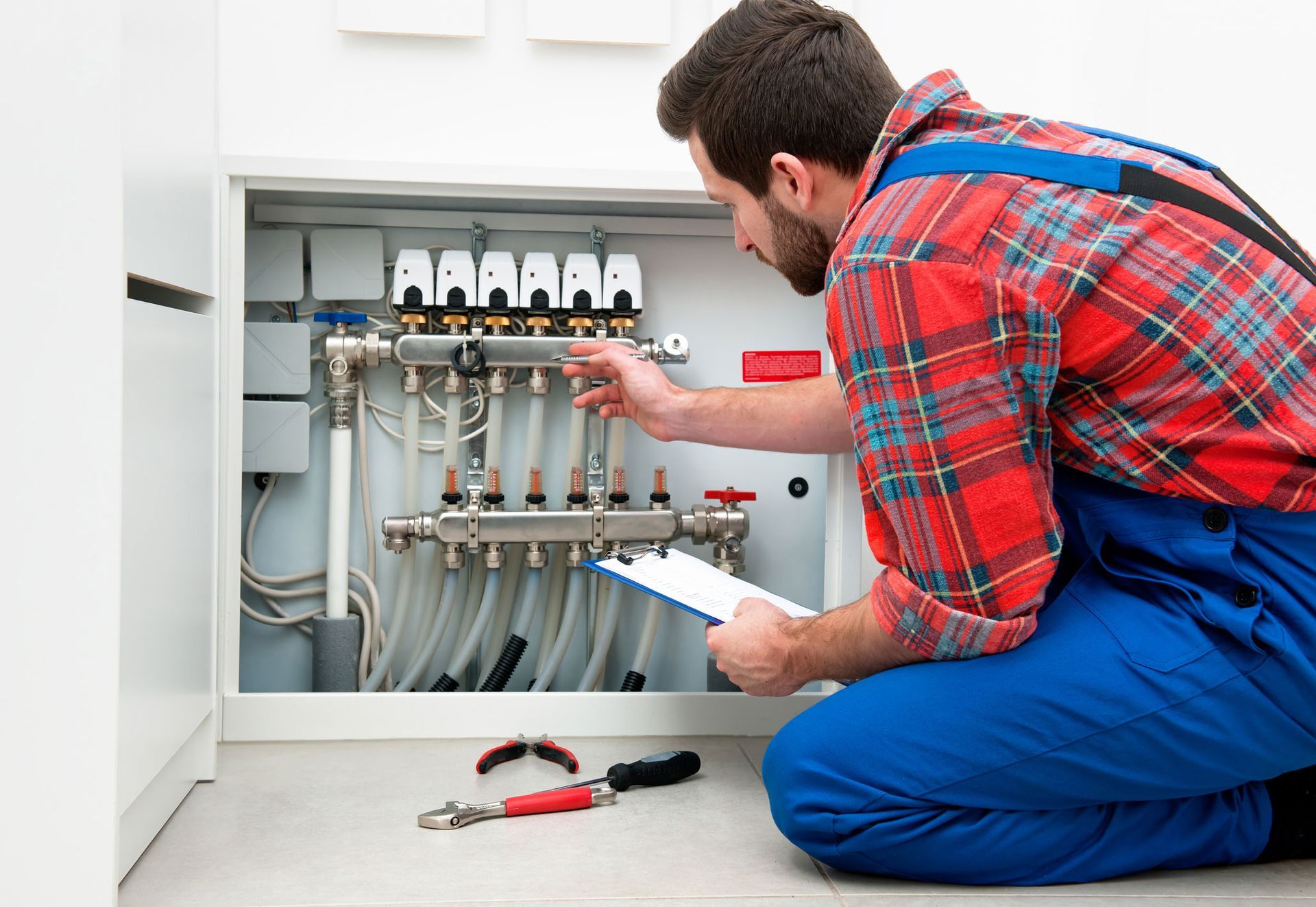 Plumber in blue overalls and plaid shirt examines pipes, holding a clipboard, in a white cabinet.