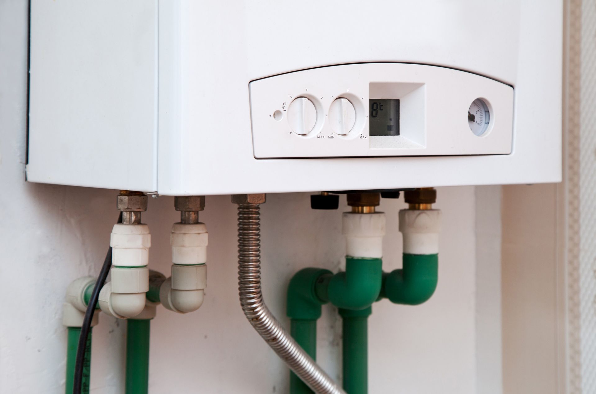 White wall-mounted gas boiler with pipes and gauges. Green and white pipes connect to the appliance.