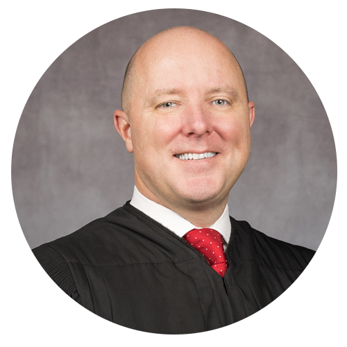 Judge in black robe, red tie, smiling in front of a gray background.