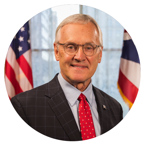 Man in suit, glasses, and red tie, smiles in front of American and Ohio flags.