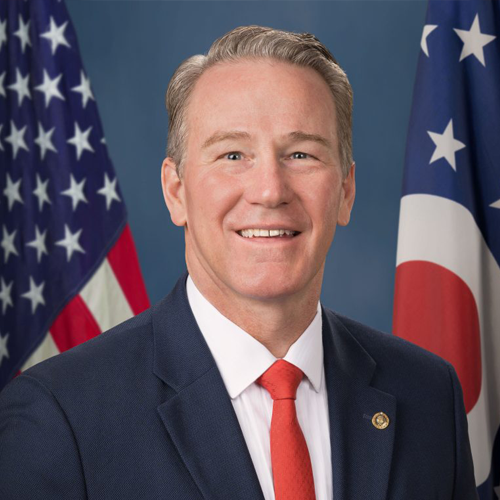 Portrait of a smiling man in a suit and tie, with American and Ohio flags in the background.