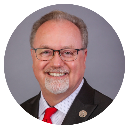 Smiling man with glasses, goatee, in a suit and red tie, against a gray backdrop.