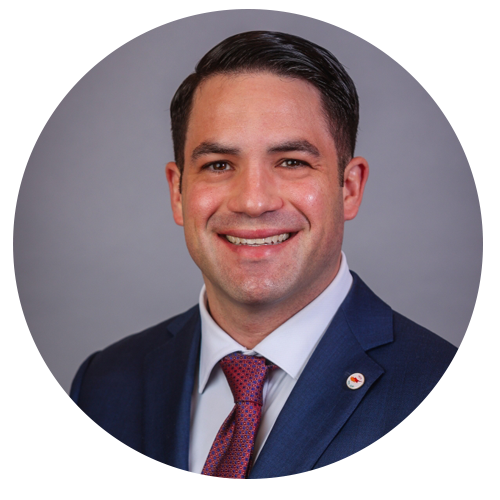 Man in a suit smiles, dark hair, red tie, oval portrait against a gray background.