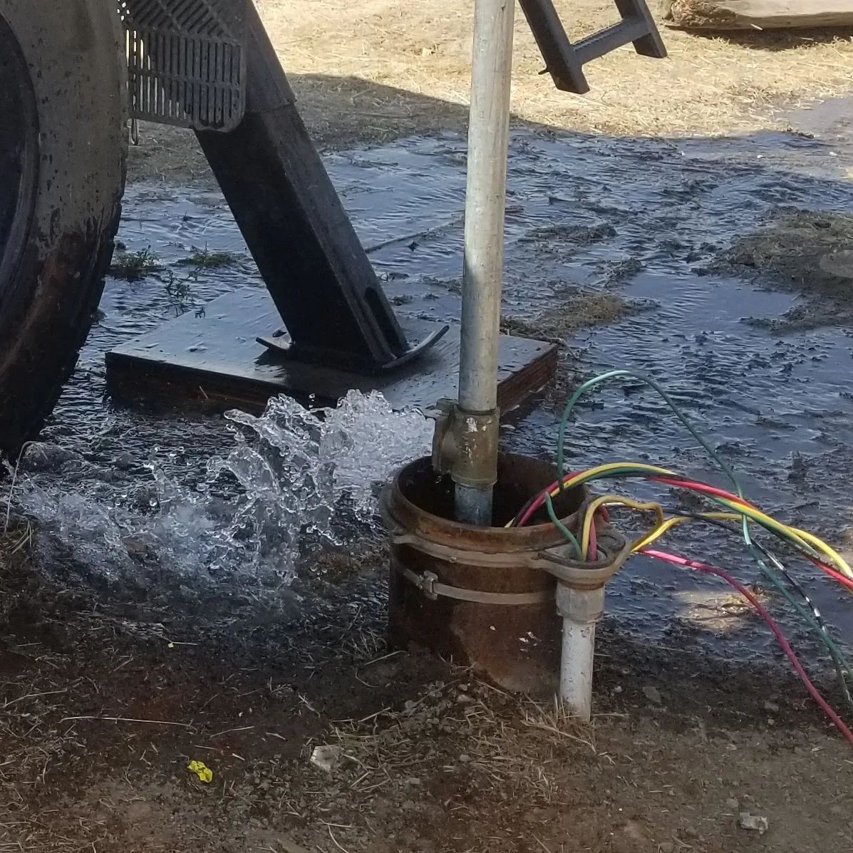 Well water gushing from a pipe, with wires connected; near a vehicle wheel and metal platform.