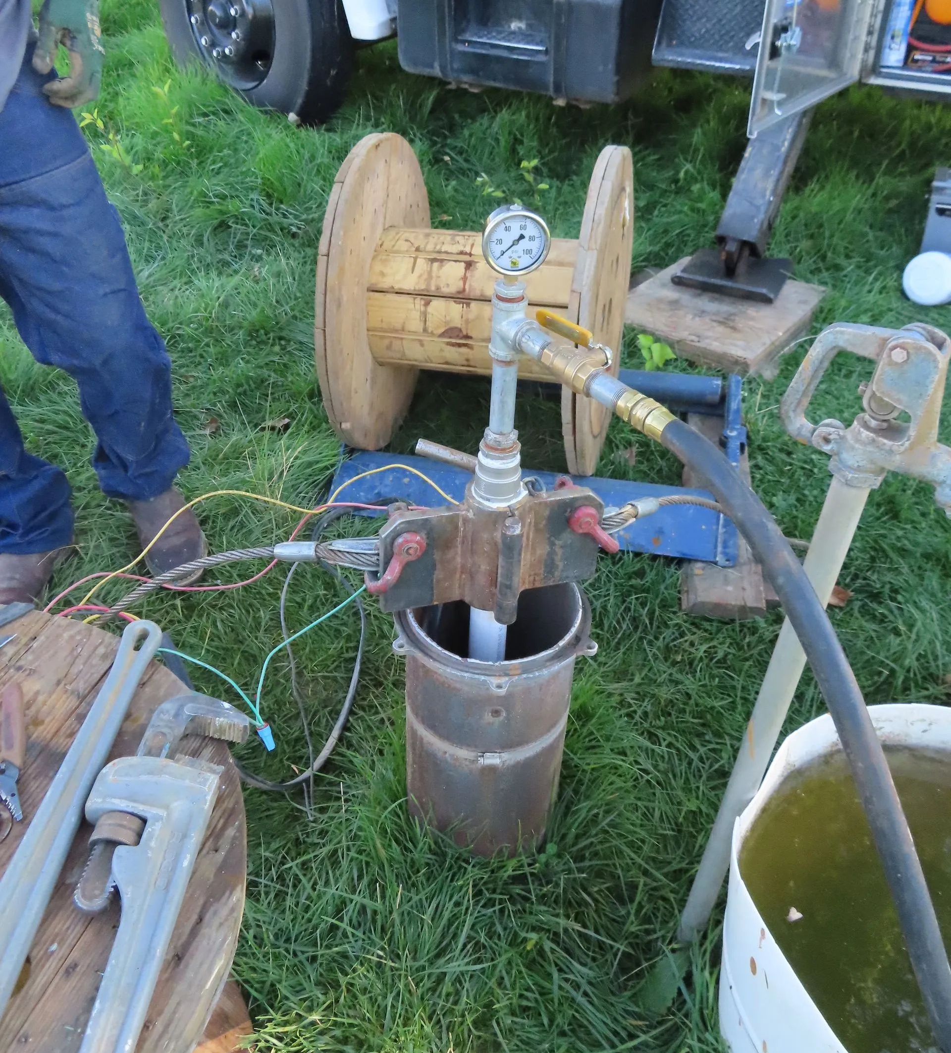 Well water testing setup with gauge, spools, pipes, and well casing in a grassy area.