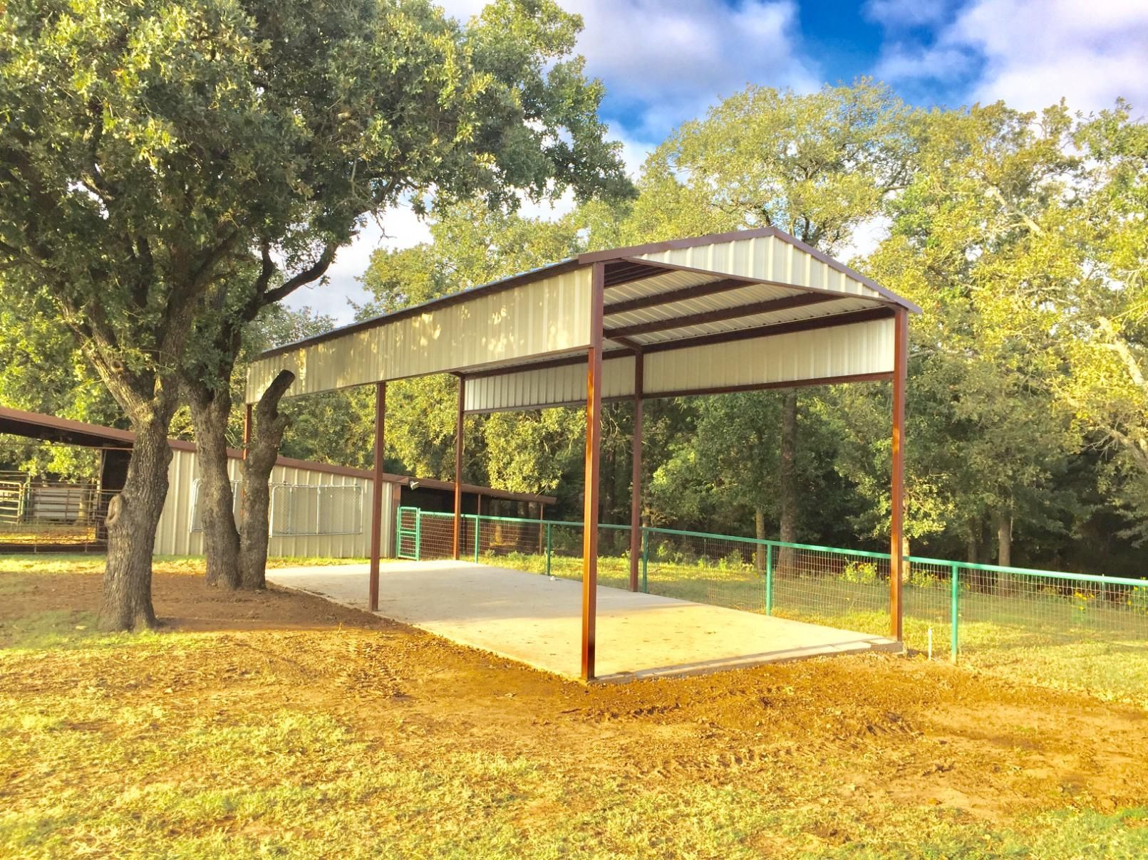 A metal-roofed carport structure with support posts standing on a concrete slab in a grassy, tree-filled outdoor area.
