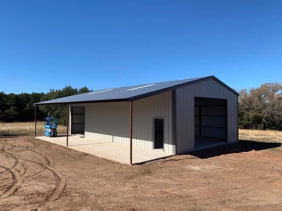A light-colored metal workshop building with an open lean-to side on a rural dirt lot under a clear blue sky.